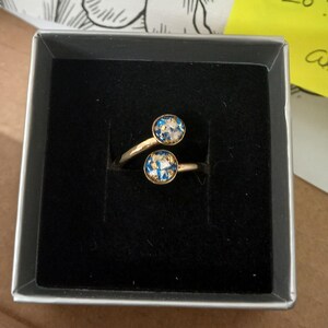 May include: A gold-toned bypass ring featuring two circular settings, each containing a mix of blue, white, and gold-colored flecks. The ring is presented inside a square box with a black velvet interior lining, showcasing the two distinct circular ends.