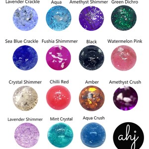 May include: A collection of fourteen circular cabochons in various colors and textures, including Lavender Crackle, Aqua, Amethyst Shimmer, Green Dichro, Sea Blue Crackle, Fushia Shimmmer, Black, Watermelon Pink, Crystal Shimmer, Chilli Red, Amber, Amethyst Crush, Lavender Shimmer, and Mint Crystal, with a black circular logo featuring the white text ahj.