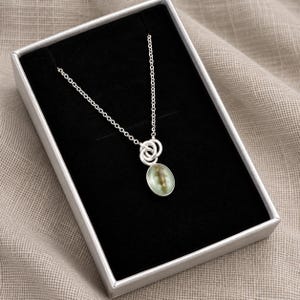 May include: A silver necklace featuring a delicate chain and a pendant with two interlocking silver rings attached to an oval-shaped charm. The charm has a smooth, light green surface with a subtle, darker organic-looking inclusion inside.