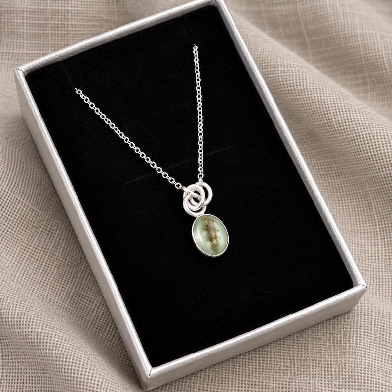 May include: A silver necklace featuring a delicate chain and a pendant with two interlocking silver rings attached to an oval-shaped charm. The charm has a smooth, light green surface with a subtle, darker organic-looking inclusion inside.