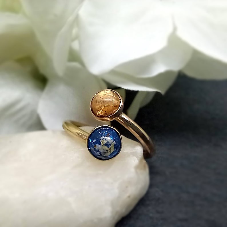 May include: A gold-toned bypass ring featuring two circular settings. One setting contains a textured amber-colored inlay, while the other setting features a deep blue inlay with embedded white flecks, creating a distinct contrast in color.