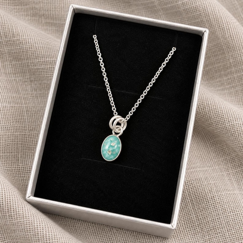 May include: A silver chain necklace featuring an oval pendant with a marbled turquoise and white resin inlay containing small embedded clear crystals. The necklace is presented inside a rectangular box with a black velvet-like interior lining.