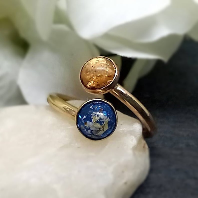 May include: A gold-toned bypass ring featuring two circular settings. One setting contains a textured amber-colored inlay, while the other contains a deep blue inlay with embedded iridescent flecks. The band is smooth and polished gold-toned metal.