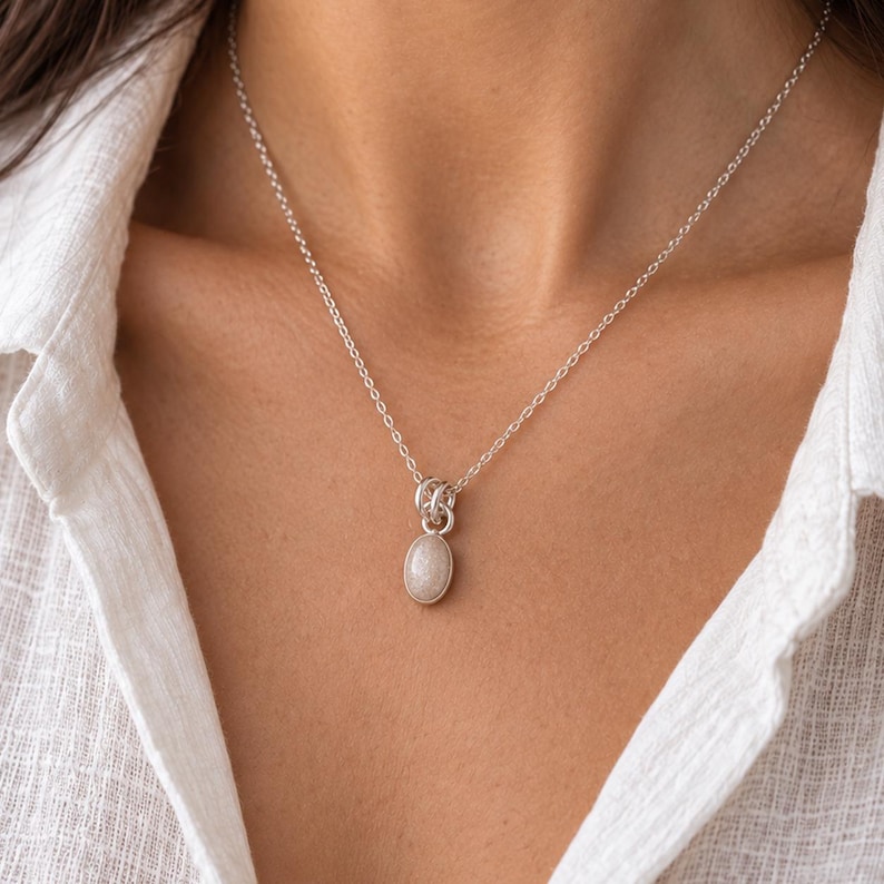 May include: A silver chain necklace featuring a small, oval-shaped pendant with a smooth, light-colored stone. The pendant is attached to the chain by a series of interconnected silver rings, creating a simple and clean design for the jewelry piece.
