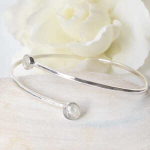 May include: A silver bypass bangle bracelet featuring a smooth, polished metal band that curves around the wrist. At each open end of the bracelet, there is a single, round, faceted clear gemstone set in a simple silver bezel setting.