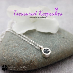 May include: Silver necklace with a small round pendant containing a black stone. The pendant is set in a silver bezel. The necklace is on a grey stone background. The text "Treasures Keepsakes memorial jewellery" is at the top of the image.