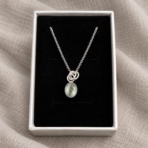 May include: A silver chain necklace featuring a small, oval-shaped pendant with a mottled, pale green and grey stone inlay. The pendant is attached to the chain by two interlocking silver rings, presented inside a rectangular box with black lining.