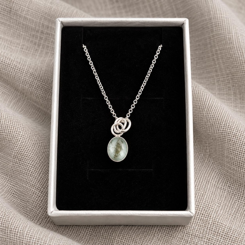 May include: A silver chain necklace featuring a small, oval-shaped pendant with a mottled, pale green and grey stone inlay. The pendant is attached to the chain by two interlocking silver rings, presented inside a rectangular box with black lining.