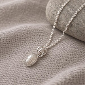 May include: A silver necklace featuring an oval pendant with a speckled, light-colored stone inlay. The pendant is attached to a delicate silver chain by two circular silver rings. The metal has a polished finish, and the stone has a textured appearance.