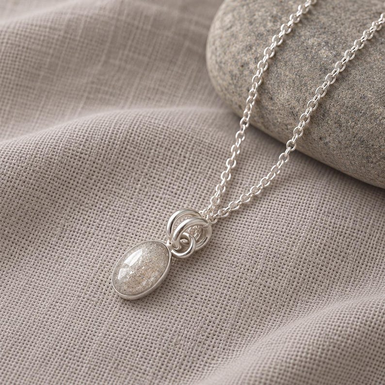 May include: A silver necklace featuring an oval pendant with a speckled, light-colored stone inlay. The pendant is attached to a delicate silver chain by two circular silver rings. The metal has a polished finish, and the stone has a textured appearance.