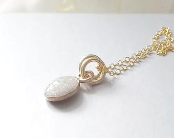May include: A gold-toned chain necklace featuring a small, oval-shaped pendant with a textured, shimmering white surface. The pendant is attached to the chain by two circular gold-toned jump rings, creating a simple and delicate jewelry piece.