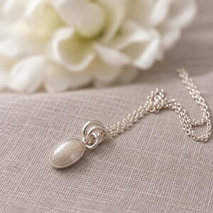 May include: A silver necklace featuring an oval pendant with a shimmering, light-colored stone inlay. The pendant is attached to a delicate silver chain by two small, circular silver rings, creating a simple and clean design for this piece of jewelry.