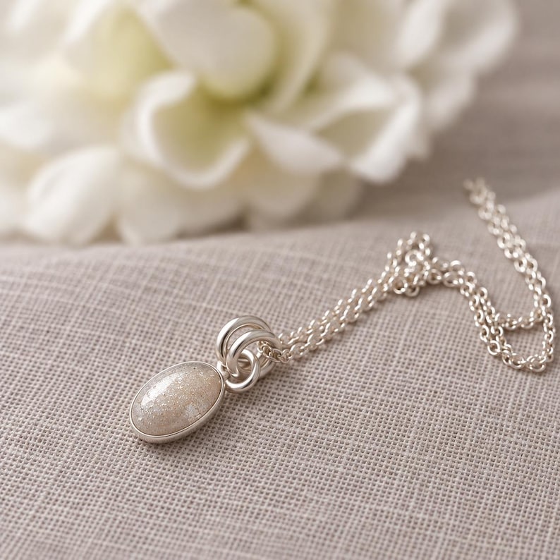 May include: A silver necklace featuring an oval pendant with a shimmering, light-colored stone inlay. The pendant is attached to a delicate silver chain by two small, circular silver rings, creating a simple and clean design for this piece of jewelry.