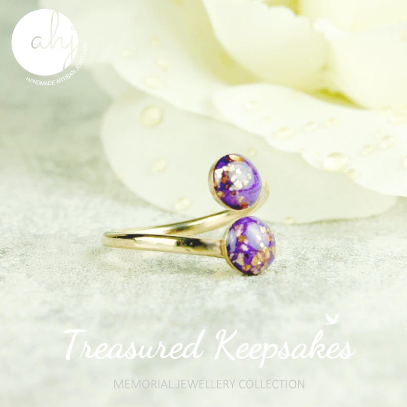 May include: A gold-toned bypass ring featuring two circular settings at each end. Each setting contains a purple resin inlay with embedded gold-colored metallic flakes. The ring has a smooth, polished metal band that curves to form an open design.
