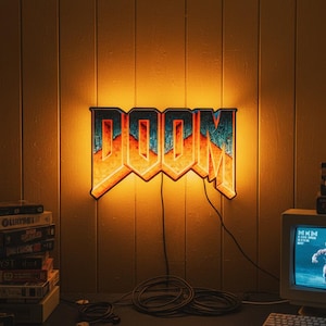 Könnte beinhalten: Beleuchtetes "DOOM"-Schild mit Retro-Videospiel-Ästhetik. Das Schild ist an einer Wand mit Holzverkleidung angebracht, mit einem Stapel Spielkassetten und einem Vintage-Computermonitor, der eine Spielszene anzeigt.