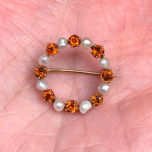 May include: A gold brooch with a circular design featuring alternating orange gemstones and white pearls.