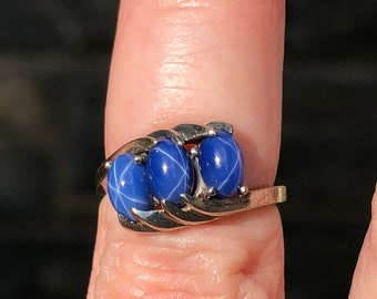 May include: A silver ring featuring three oval, blue star sapphires. The gemstones are set in a decorative, swirling silver band. The ring is displayed on a finger, showcasing the polished stones and the ring's design.