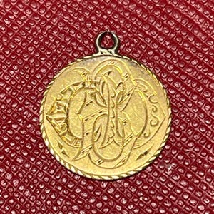 Puede incluir: Un colgante dorado con un diseño ornamentado, posiblemente un monograma o emblema. Tiene forma circular con un borde texturizado y un pequeño lazo para una cadena. El fondo es una superficie roja oscura texturizada.