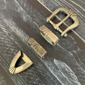 May include: A four-piece metal belt hardware set in a muted brass finish. The set includes a rectangular buckle with a prong, two rectangular belt loops, and a triangular belt tip, each featuring a raised fan-shaped decorative detail.