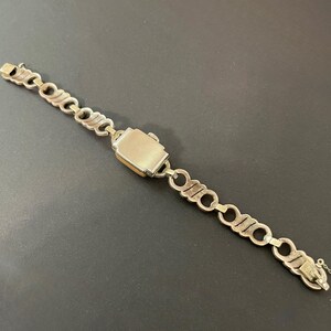 Pu&ograve; includere: Orologio da polso vintage color argento con quadrante rettangolare e bracciale a maglie. Il quadrante dell'orologio ha un bordo color oro. Il bracciale presenta una serie di maglie interconnesse e stilizzate. L'orologio &egrave; su una superficie scura.