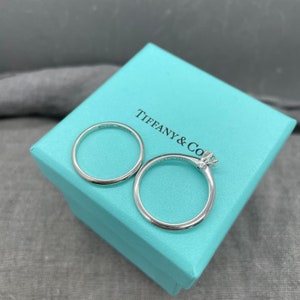 May include: Two silver wedding bands in a Tiffany & Co. blue box. The band on the left is a plain band, and the band on the right has a small diamond set in the center.