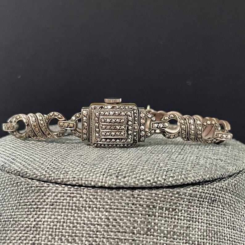 Pu&ograve; includere: Un orologio vintage color argento con quadrante quadrato e cinturino decorativo. Il quadrante dell'orologio &egrave; incrostato di piccole pietre scintillanti e il cinturino presenta maglie ornate. L'orologio &egrave; esposto su una superficie grigia su sfondo scuro.