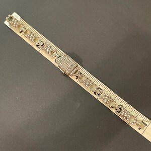 Pu&ograve; includere: Bracciale argentato con chiusura rettangolare e maglie decorative, impreziosito da piccoli strass trasparenti. Il bracciale &egrave; lungo circa 17.78 cm, mostrato accanto a un righello su una superficie scura.