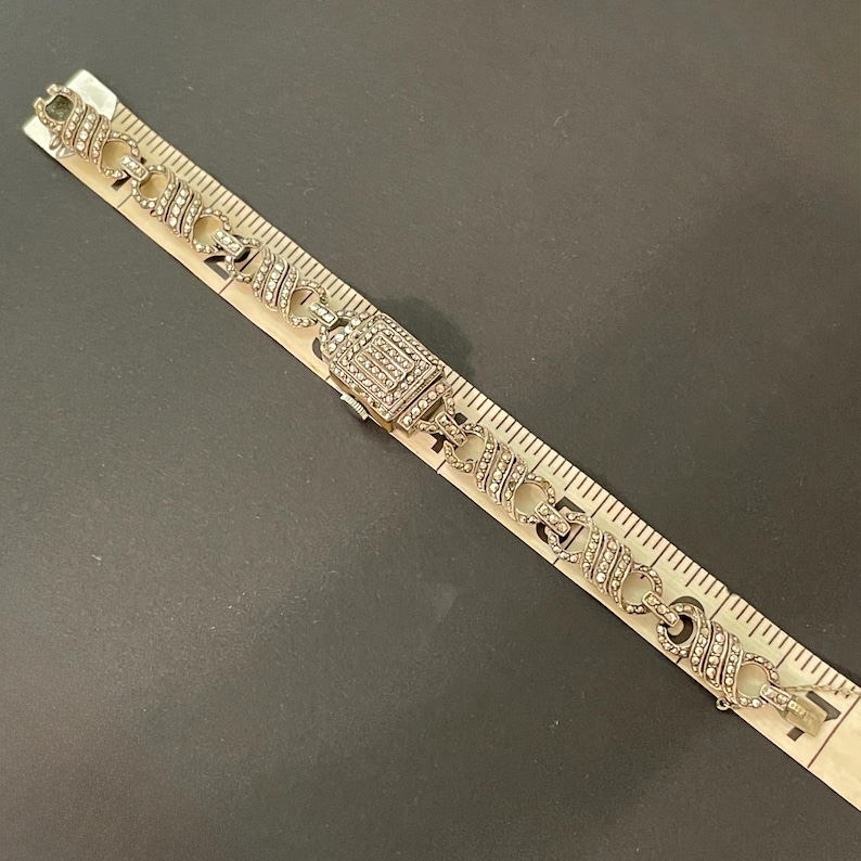 Pu&ograve; includere: Bracciale argentato con chiusura rettangolare e maglie decorative, impreziosito da piccoli strass trasparenti. Il bracciale &egrave; lungo circa 17.78 cm, mostrato accanto a un righello su una superficie scura.