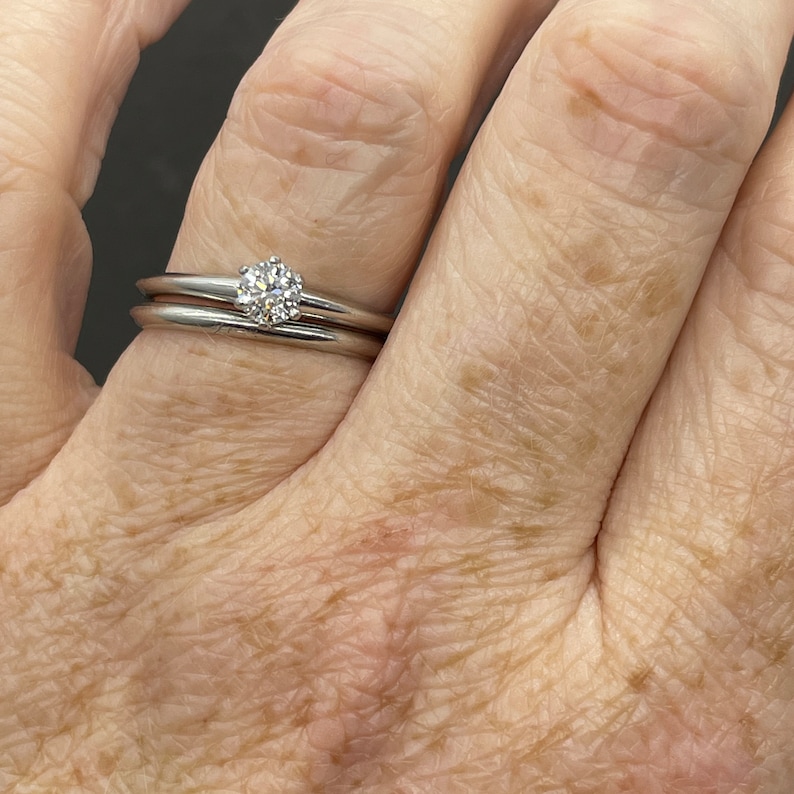 May include: A close-up of a hand wearing a simple silver wedding band and a solitaire engagement ring with a round diamond.