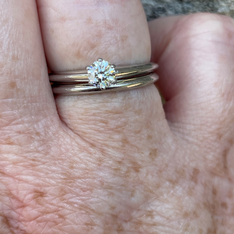 May include: A close-up of a hand wearing two silver bands. The top band features a single, round, clear diamond.