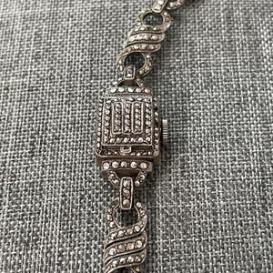 Pu&ograve; includere: Primo piano di un bracciale per orologio vintage argentato. Il quadrante dell'orologio &egrave; rettangolare con un design dettagliato e ornato. I collegamenti del bracciale presentano un design a spirale con piccoli abbellimenti scintillanti.