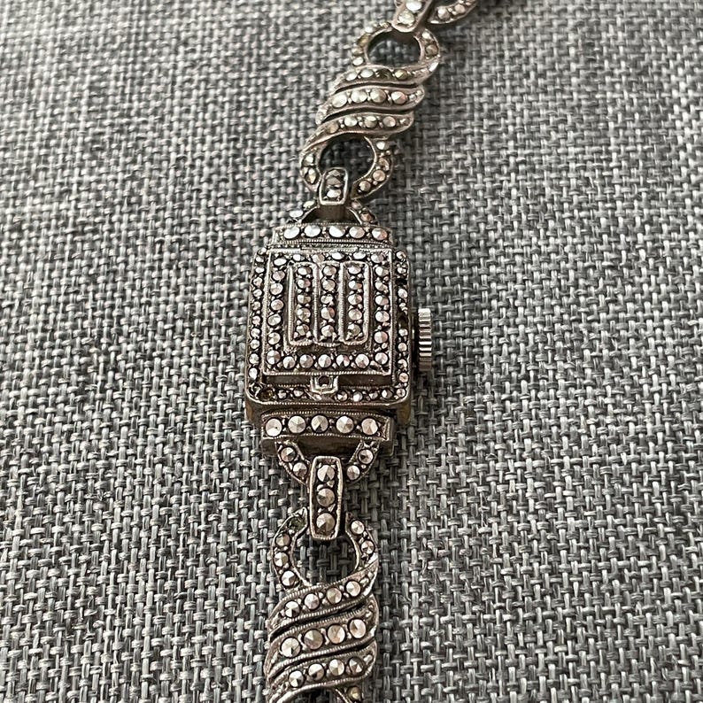 Pu&ograve; includere: Primo piano di un bracciale per orologio vintage argentato. Il quadrante dell'orologio &egrave; rettangolare con un design dettagliato e ornato. I collegamenti del bracciale presentano un design a spirale con piccoli abbellimenti scintillanti.