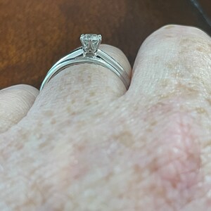 May include: A close-up of a silver engagement ring with a single round diamond on a person's finger.