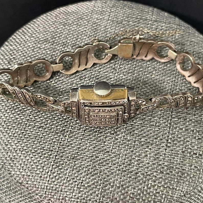 Pu&ograve; includere: Un orologio da polso vintage color argento con quadrante rettangolare e cinturino decorativo. Il quadrante dell'orologio &egrave; dorato con una corona argentata. Il cinturino presenta una serie di maglie decorative ed &egrave; incrostato di piccole pietre scintillanti.