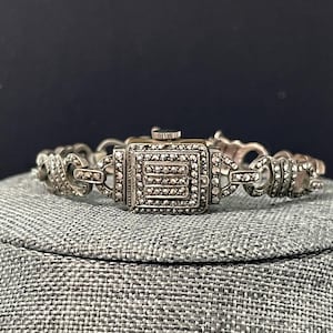 Pu&ograve; includere: Orologio vintage color argento con quadrante quadrato e dettagli in marcasite. L'orologio presenta un bracciale a maglie decorativo. Il quadrante dell'orologio ha una forma rettangolare con una piccola corona. L'orologio &egrave; esposto su una superficie grigia.