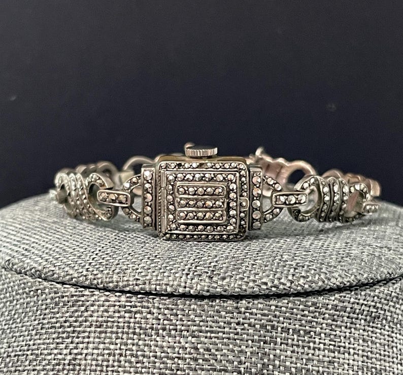 Pu&ograve; includere: Orologio vintage color argento con quadrante quadrato e dettagli in marcasite. L'orologio presenta un bracciale a maglie decorativo. Il quadrante dell'orologio ha una forma rettangolare con una piccola corona. L'orologio &egrave; esposto su una superficie grigia.