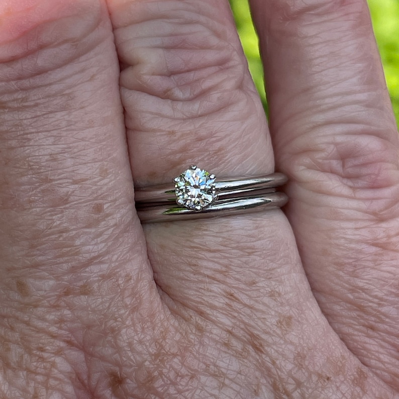 May include: A close-up of a silver ring with a single round diamond set in the center. The ring is worn on the finger of a person's hand.