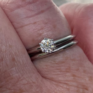 May include: A close-up of a silver ring with a single round diamond set in the center. The ring is worn on a finger with a second silver band worn underneath.