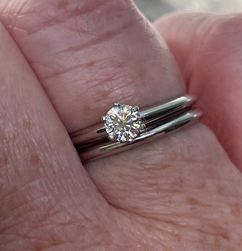 May include: A close-up of a silver ring with a single round diamond set in the center. The ring is worn on a finger with a second silver band worn underneath.