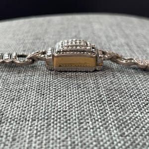 Pu&ograve; includere: Primo piano di un braccialetto argentato con una chiusura rettangolare dorata. La chiusura &egrave; incastonata con piccole pietre preziose trasparenti. Il braccialetto ha un design delicato e ornato.