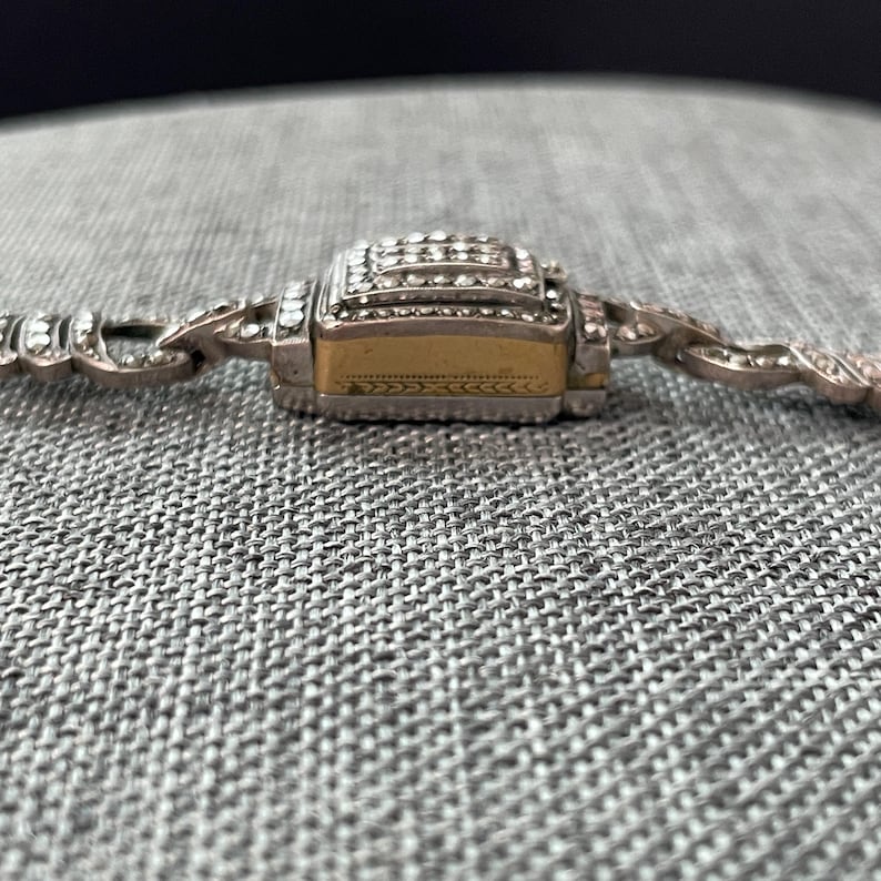 Pu&ograve; includere: Primo piano di un braccialetto argentato con una chiusura rettangolare dorata. La chiusura &egrave; incastonata con piccole pietre preziose trasparenti. Il braccialetto ha un design delicato e ornato.