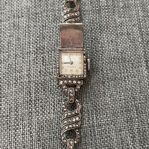 Pu&ograve; includere: Orologio da polso vintage color argento con quadrante rettangolare e coperchio incernierato. Il quadrante ha uno sfondo color crema con numeri e lancette nere. Il cinturino &egrave; una catena delicata con accenti decorativi.