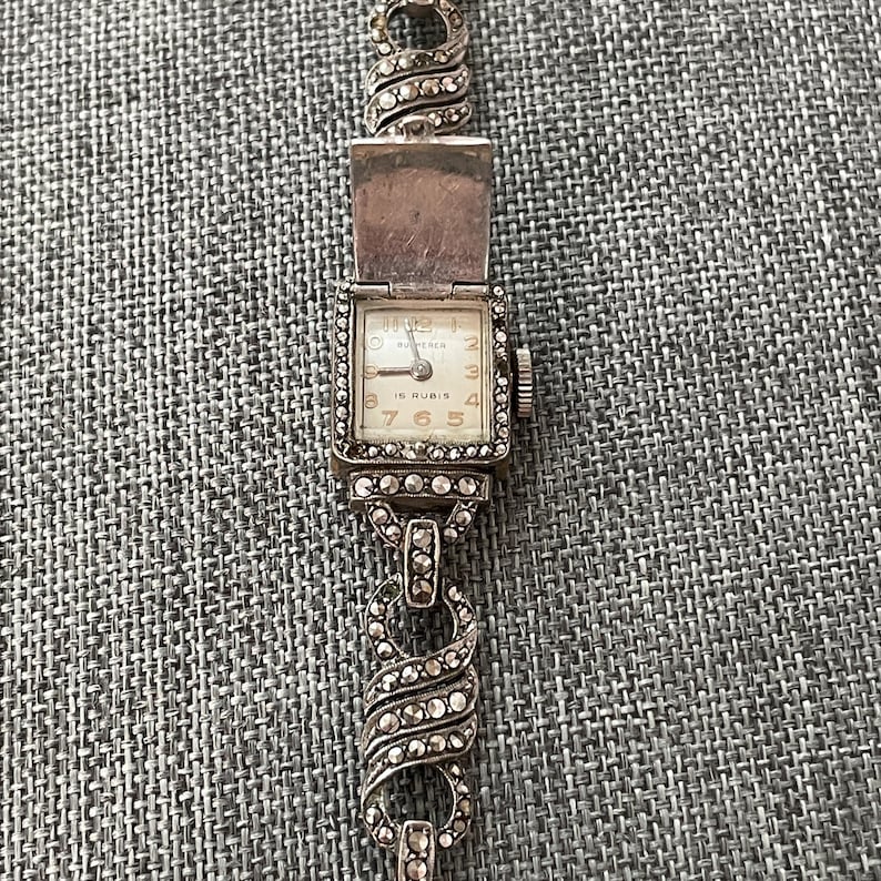 Pu&ograve; includere: Orologio da polso vintage color argento con quadrante rettangolare e coperchio incernierato. Il quadrante ha uno sfondo color crema con numeri e lancette nere. Il cinturino &egrave; una catena delicata con accenti decorativi.