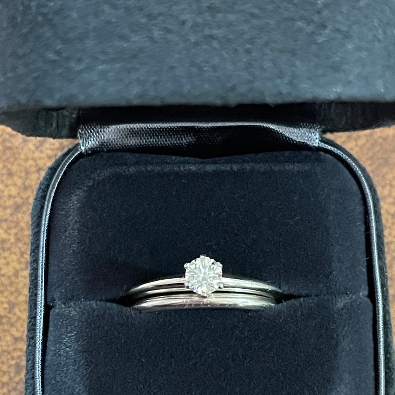 May include: A set of two silver wedding bands with a single diamond solitaire ring in the center. The rings are in a black velvet ring box.