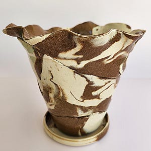 May include: A handcrafted ceramic planter with a unique, wavy rim. The planter features a marbled design in shades of brown and cream, with a matching saucer. The textured surface adds to its artistic appeal. Ideal for indoor plants.