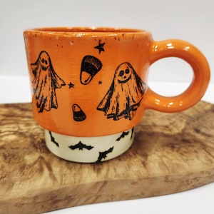 May include: A ceramic mug with a glossy orange upper section featuring black ink illustrations of two ghosts, candy corn, and stars. The base is a contrasting cream color decorated with a repeating pattern of small black flying bats.