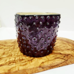 May include: A small, textured, purple ceramic pot with a bumpy surface. The pot has a dark interior and is set on a wooden cutting board. The pot is a deep purple color with small, dark, round bumps covering the exterior.