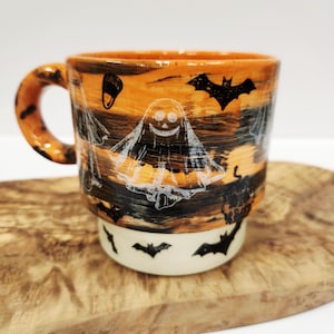 May include: A ceramic mug with a handle, featuring a horizontal orange and black striped pattern. The mug is decorated with white line drawings of ghosts and black silhouettes of bats. The base of the mug is a solid cream color with black bat silhouettes.