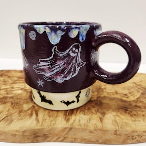 May include: A ceramic mug with a dark purple upper section featuring a white ghost and star design, and a cream-colored base decorated with black bat silhouettes. The mug has a thick, rounded handle and a glossy finish throughout the exterior.
