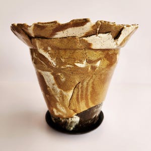 May include: A handcrafted ceramic vase with a unique, textured design. The vase features a flared rim and a marbled pattern in shades of brown, tan, and white. It sits on a small, round, black base. A decorative piece for home decor.